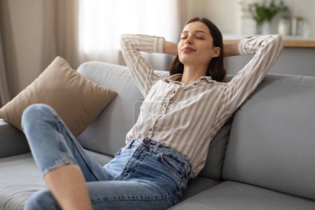 Photo for Tranquil young woman enjoying peaceful moment, reclining with hands behind head on soft sofa, embodying relaxation and comfort at home, free space - Royalty Free Image