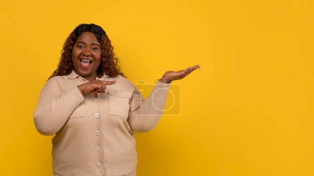 Photo for Excited young overweight black woman wearing beige shirt holding and pointing at something invisible on her palm hand, showing nice product, yellow background, copy space - Royalty Free Image