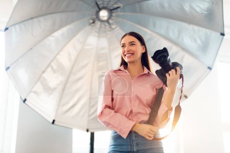 Téléchargez les photos : Jeune photographe heureuse tenant un appareil photo reflex numérique professionnel, debout sous un équipement d'éclairage avec parapluie réfléchissant dans un studio photo - en image libre de droit
