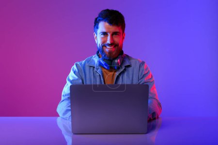 Téléchargez les photos : Professionnel en ligne. programmeur caucasien gars assis avec ordinateur portable au bureau, regardant la caméra, travaillant à l'ordinateur en violet et bleu studio néon. Carrières sur Internet, possibilités d'apprentissage en ligne - en image libre de droit