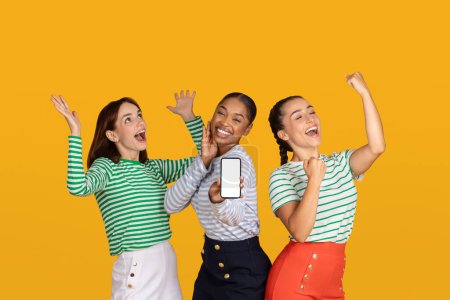 Photo for Emotional young ladies girlfriends showing smartphone with white blank screen mockup copy space gesturing and exclaiming, celebrating beginning of season sale, yellow background - Royalty Free Image