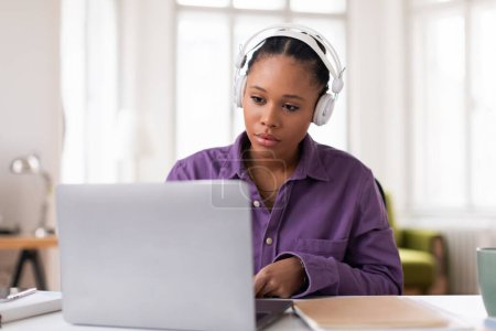 Konzentrierte junge Frau, die Kopfhörer trägt, während sie an ihrem Laptop arbeitet, in lila Hemd, konzentriert auf ihre E-Learning-Aufgabe zu Hause