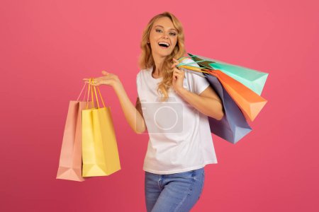 Photo for Seasonal Shopper. Beautiful European blonde lady carrying shopping bags and expressing happiness over great sales, standing with purchases in studio setting with pink background. Discount promotion - Royalty Free Image