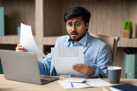 Foto de Empresario hindú infeliz manejando problemas de papeleo leyendo informes de negocios negativos y correspondencia, sentado en su escritorio frente a la computadora portátil, se enfrenta a desafíos de trabajo en el entorno de la oficina - Imagen libre de derechos