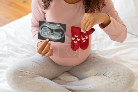 Photo for Young pregnant woman holding sonogram next to tiny red heart-patterned baby socks, unrecognizable african american expectant mother celebrating her journey to motherhood, sitting on bed at home - Royalty Free Image