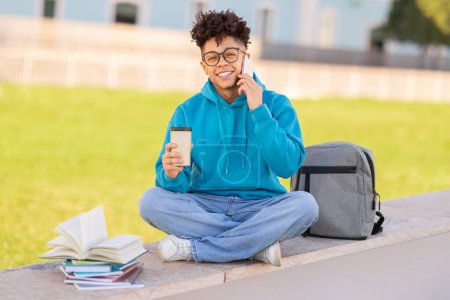 Glücklicher schwarzer Student, der telefoniert und mit Imbisskaffee im Freien auf dem Campus der Universität sitzt, in der Pause kommuniziert und mit Büchern und Rucksack in die Kamera lächelt