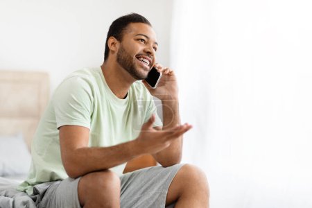 Téléchargez les photos : Joyeux beau jeune homme afro-américain assis sur le lit le matin, parlant au téléphone à la maison, avoir une conversation avec l'amant tout en appréciant week-end, en regardant l'espace de copie - en image libre de droit