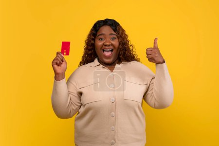 Foto de Feliz gordita millennial africana americana mujer con el pelo largo rizado mostrando tarjeta de crédito de plástico rojo y pulgar hacia arriba, recomendando su banco, aislado sobre fondo amarillo - Imagen libre de derechos