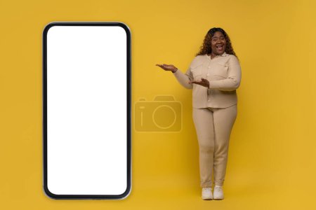 Photo for Positive chubby long-haired millennial african american lady wearing beige costume pointing at huge phone with white blank screen mockup copy space on yellow background, showing online offer - Royalty Free Image