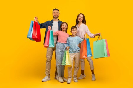 Photo for Family Shopping. Joyful Young Parents And Their Kids Holding Bright Shopper Bags And Having Fun Together While Standing On Yellow Background, Cheerful Mom, Dad And Children Enjoying Seasonal Sales - Royalty Free Image