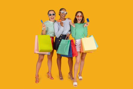 Foto de Tres damas jóvenes multirraciales con gafas de sol y trajes de lujo disfrutando de compras, llevando coloridas compras de bolsas de papel y mostrando tarjetas de crédito, fondo de estudio amarillo, longitud completa - Imagen libre de derechos