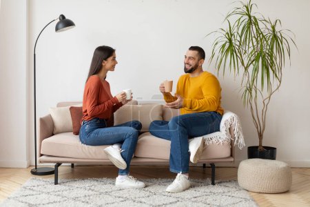 Photo for Happy Young Spouses Chatting And Drinking Coffee Together At Home, Loving Millennial Couple Relaxing On Comfortable Couch In Living Room, Talking And Smiling, Enjoying Time With Each Other - Royalty Free Image