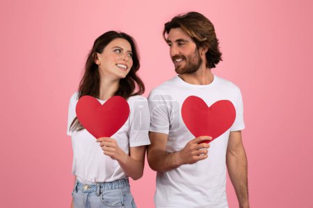 Foto de Mujer y hombre europeos sonrientes sosteniendo cada uno un corazón de papel rojo sobre sus camisetas blancas, mirándose con afecto, simbolizando el amor y el día de San Valentín sobre un fondo rosa - Imagen libre de derechos