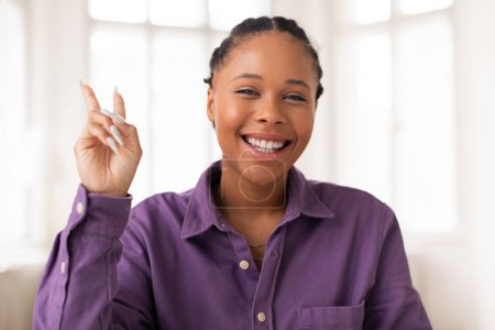 Photo for Cheerful black teenage girl wearing casual purple shirt smiling and gesturing peace sign with her hand, exuding relaxed and joyful demeanor - Royalty Free Image
