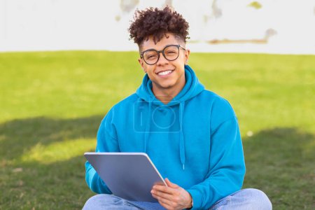 Téléchargez les photos : Toothy étudiant noir gars en lunettes surfer sur Internet sur tablette numérique, assis à l'extérieur au parc vert, souriant à la caméra. Portrait de l'apprenant en ligne réussie avec gadget - en image libre de droit