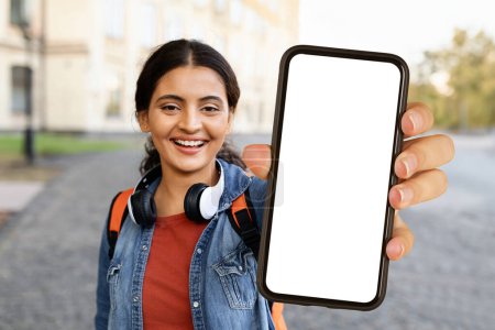 Photo for Cheerful pretty young eastern lady student with wireless headphones and backpack showing phone with white empty screen, recommending nice mobile app, walking by campus, mockup - Royalty Free Image