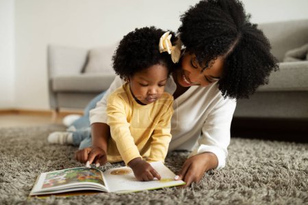 Photo for Parenthood concept. Adorable curly little african american girl playing with her mother, happy black toddler and mom reading book on floor, home interior, closeup, copy space - Royalty Free Image
