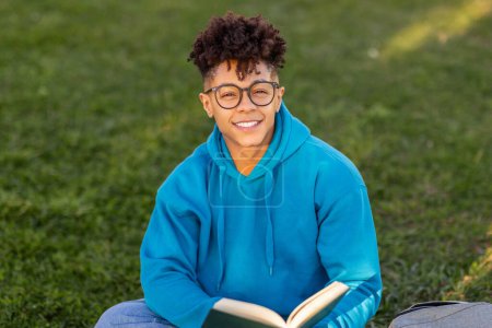 Photo for Brazilian student guy wearing glasses and happily holding book relaxing in park outside, smiling to camera, enjoying reading and educational leisure outside university campus - Royalty Free Image