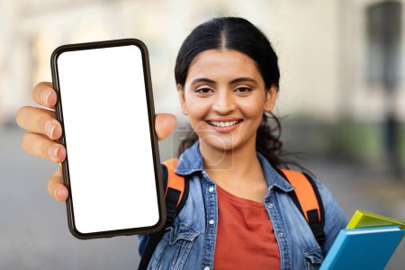 Photo for Smiling young indian woman student with textbooks in her hand showing phone with white empty screen mockup copy space, recommending educational app, university campus background - Royalty Free Image