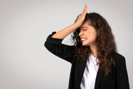 Businesswoman with curly hair showing facepalm expression of frustration or embarrassment, wearing smart suit on light grey background, copy space