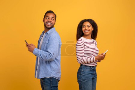 Photo for Cheerful young couple holding smartphone and glancing back over their shoulders, smiling african american man and woman advertising dating app, standing on yellow studio background, copy space - Royalty Free Image