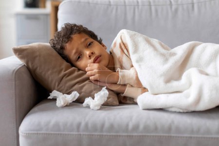 Photo for Ill little black boy resting under blanket on couch at home, with tissues scattered around him, sick preteen male kid looking upset and tired while resting on sofa in living room interior - Royalty Free Image