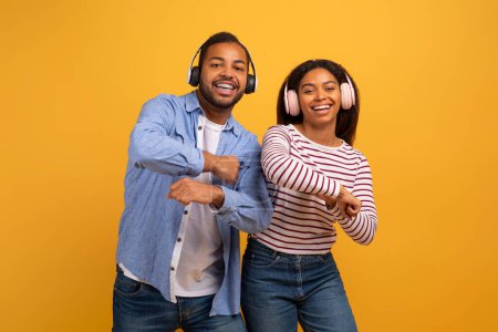 Foto de Sonriendo joven pareja negra bailando y disfrutando de la música con auriculares de moda, feliz millennial afroamericano hombre y mujer mirando a la cámara con expresión alegre, divertirse en el fondo amarillo - Imagen libre de derechos