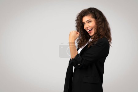 Foto de Encantada joven empresaria en traje formal celebra con la bomba de puño, mostrando expresión alegre y entusiasmo, sobre fondo gris, espacio libre - Imagen libre de derechos