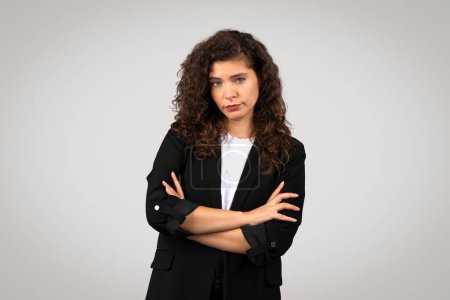 Skeptical young businesswoman with curly hair and black blazer stands with arms crossed, looking serious or uncertain, against neutral grey background