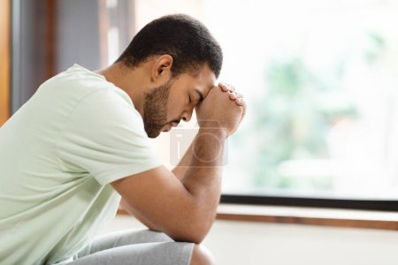 Photo for Closeup of unhappy millennial african american guy suffering from loneliness. Upset young black man wearing pajamas sitting on couch next to window at home, feeling depressed, side view, copy space - Royalty Free Image