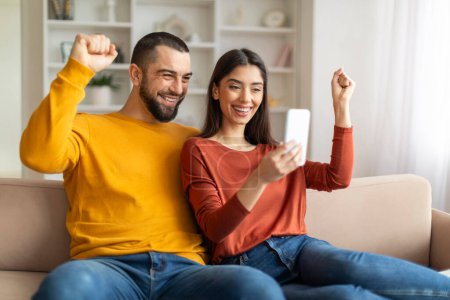 Photo for Happy Young Couple Celebrating Success While Looking At Smartphone Screen At Home, Joyful Millennial Spouses Cheering Win, Emotionally Reacting To Good News, Raising Fists With Excitement - Royalty Free Image