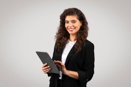 Foto de Joven empresaria confiada con el pelo rizado sonriendo y sosteniendo la tableta digital, que representa profesionalismo moderno y tecnología en los negocios - Imagen libre de derechos