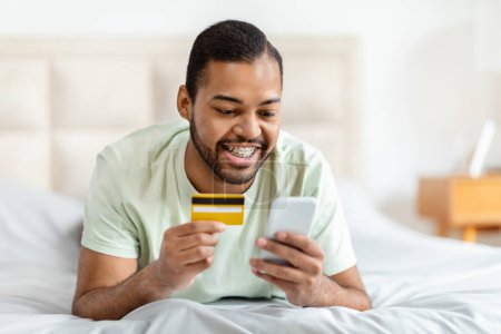Photo for E-commerce, retail, virtual banking. Happy handsome african american man lying on bed, using smartphone and plastic bank card, making online order while staying home at weekend, copy space - Royalty Free Image