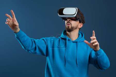 Photo for Engrossed man wearing virtual reality headset and gesturing with his hands, amazed millennial guy interacting with digital world, enjoying modern technologies, standing against blue studio background - Royalty Free Image