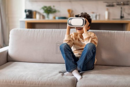 Photo for Excited little black boy wearing virtual reality headset sitting on couch at home, african american male kid laughing and enjoying immersive digital experience, relaxing in bright living room - Royalty Free Image