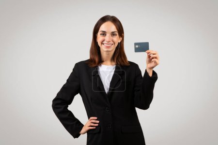 Foto de Mujer profesional caucásica milenaria con confianza en un traje negro de negocios que muestra orgullosamente una tarjeta de crédito, que representa los servicios financieros, la banca o el poder adquisitivo del consumidor. - Imagen libre de derechos