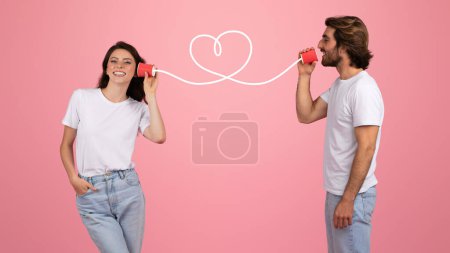 Photo for Smiling european woman and man communicating through tin can telephones with a wire shaped like a heart, symbolizing romantic connection, both in white t-shirts on a pink background - Royalty Free Image