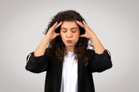 Photo for Anxious businesswoman in black blazer looking stressed with her hands on her temples, eyes closed, signaling headache or pressure, against grey background - Royalty Free Image