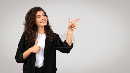 Photo for Smiling young businesswoman giving thumbs up and pointing to the side at copy space, lady dressed in black blazer and white shirt on grey background, panorama - Royalty Free Image