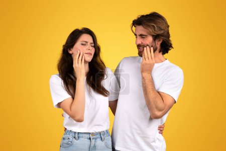 Photo for A concerned european young woman and a man touch their cheeks with pained expressions, as if experiencing toothache, both dressed in white t-shirts on a yellow background - Royalty Free Image