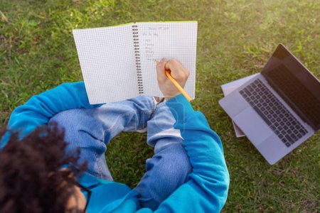 Téléchargez les photos : Vue arrière d'un étudiant noir écrivant son programme scolaire assis avec un ordinateur portable sur l'herbe, étudiant en plein air au parc du campus universitaire, planifiant des leçons en cahier d'exercices, grand angle - en image libre de droit