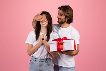 Photo for Delighted european woman with eyes covered by a mans hand, receiving a surprise gift box with a red ribbon, both wearing white t-shirts, sharing a joyful moment on a pink background - Royalty Free Image