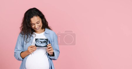 Téléchargez les photos : Joyeux jeune femme enceinte brune avec gros ventre montrant l'image échographique de son bébé, tenant la photo du foetus sur sa poitrine, fond de studio rose, panorama avec espace de copie - en image libre de droit