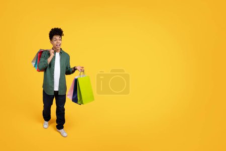 Photo for Excited young african american guy with colorful paper bags purchases walking on yellow studio background, looking at copy space and exclaiming. Shopping on season sale - Royalty Free Image