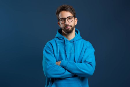 Man wearing glasses and blue hoodie looking disinterested, standing with arms crossed and slightly downcast gaze, embodying sense of boredom or disappointment, posing against dark background
