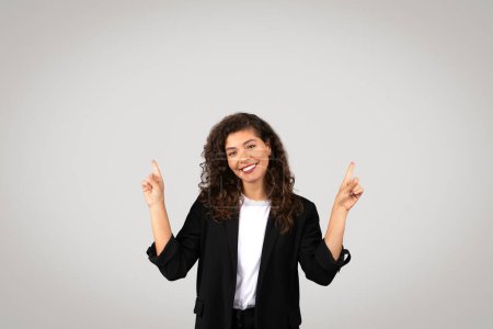 Foto de Mujer de negocios rizada sonriente en blazer negro apuntando hacia arriba al espacio libre, perfecto para anuncios de negocios, contra fondo neutro - Imagen libre de derechos