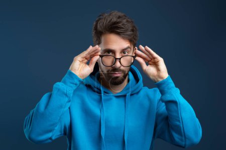 Photo for Young man adjusting his glasses with both hands and looking at camera with attentive gaze, handsome millennial guy wearing blue hoodie standing against dark studio background, closeup shot - Royalty Free Image