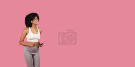 Téléchargez les photos : Jeune femme afro-américaine sportive avec des écouteurs, portant un soutien-gorge de sport blanc, tenant smartphone et regardant loin l'espace de copie sur fond rose - en image libre de droit