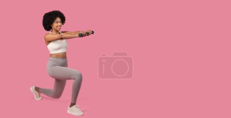 Téléchargez les photos : Femme noire joyeuse effectuant fente de remise en forme avec bras étendus tenant des haltères, portant des vêtements de sport sur fond rose, panorama avec espace libre - en image libre de droit