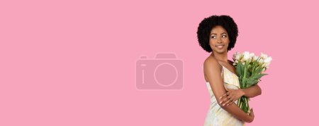Téléchargez les photos : Femme afro-américaine souriante et confiante aux cheveux bouclés, tenant un bouquet de fleurs blanches, regarde au loin sur un fond rose pastel, studio, panorama - en image libre de droit